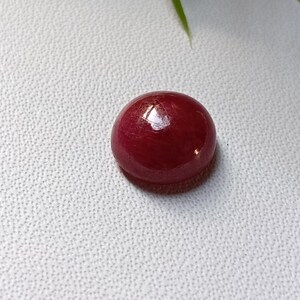 May include: A round, polished, deep red gemstone, possibly a ruby, sits on a textured white surface. The gem reflects light, showcasing its smooth, glossy surface. The overall composition is simple, highlighting the gem's color and form.