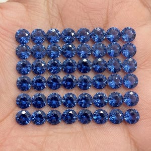 Puede incluir: Una colección de cuarenta y dos piedras preciosas azules redondas y facetadas dispuestas en un patrón de cuadrícula. Cada piedra presenta un corte brillante con múltiples facetas que reflejan la luz, mostrando un color azul profundo.