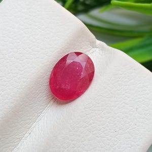Può includere: Una gemma di rubino sfaccettata, di forma ovale. La gemma è di colore rosso intenso e si trova su una superficie bianca. La pietra preziosa è il punto focale dell'immagine, con uno sfondo verde sfocato.