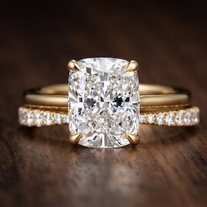 May include: A gold engagement ring set featuring a large cushion-cut centre stone held by four prongs, paired with a matching gold wedding band encrusted with a row of small round-cut diamonds along the front half of the band's visible surface.