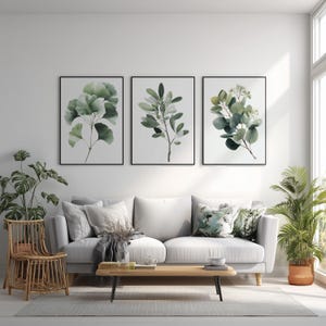Könnte beinhalten: Drei gerahmte botanische Drucke mit Aquarellillustrationen von grünen Blättern und Zweigen. Die Drucke hängen über einem weißen Sofa mit dekorativen Kissen. Ein Holztisch und Topfpflanzen vervollständigen die Wohnzimmerszene.