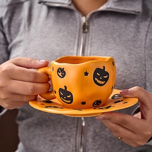 Può includere: Set tazza e piattino in ceramica arancione a tema Halloween. La tazza e il piattino sono decorati con facce di jack-o'-lantern nere e stelle. La tazza ha un manico ed è tenuta da una persona che indossa una giacca grigia con cerniera.