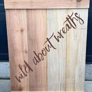 May include: Wooden wreath sign with the text "wild about wreaths" in a cursive font. The sign is made of various shades of wood and has a black hook at the top for hanging. The sign is rectangular in shape.