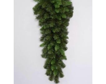May include: A dark green artificial hanging plant, resembling a cascading evergreen branch. The plant is densely packed with needle-like leaves, creating a full and textured appearance. It is designed for wall decor and adds a touch of nature to any space.