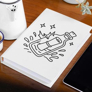 May include: A white notebook with a black line drawing of a message in a bottle. A small white mug with a blue rim and handle holds coffee. A white kettle and a tablet are also on the wooden surface.