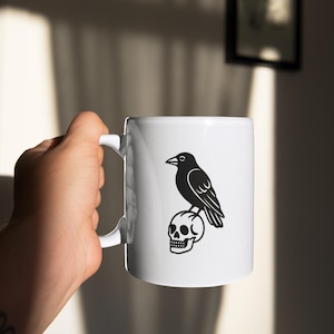 May include: White ceramic mug featuring a black illustration of a raven perched atop a skull. The mug has a curved handle and is held by a person. The design is a simple, yet striking, black and white graphic.