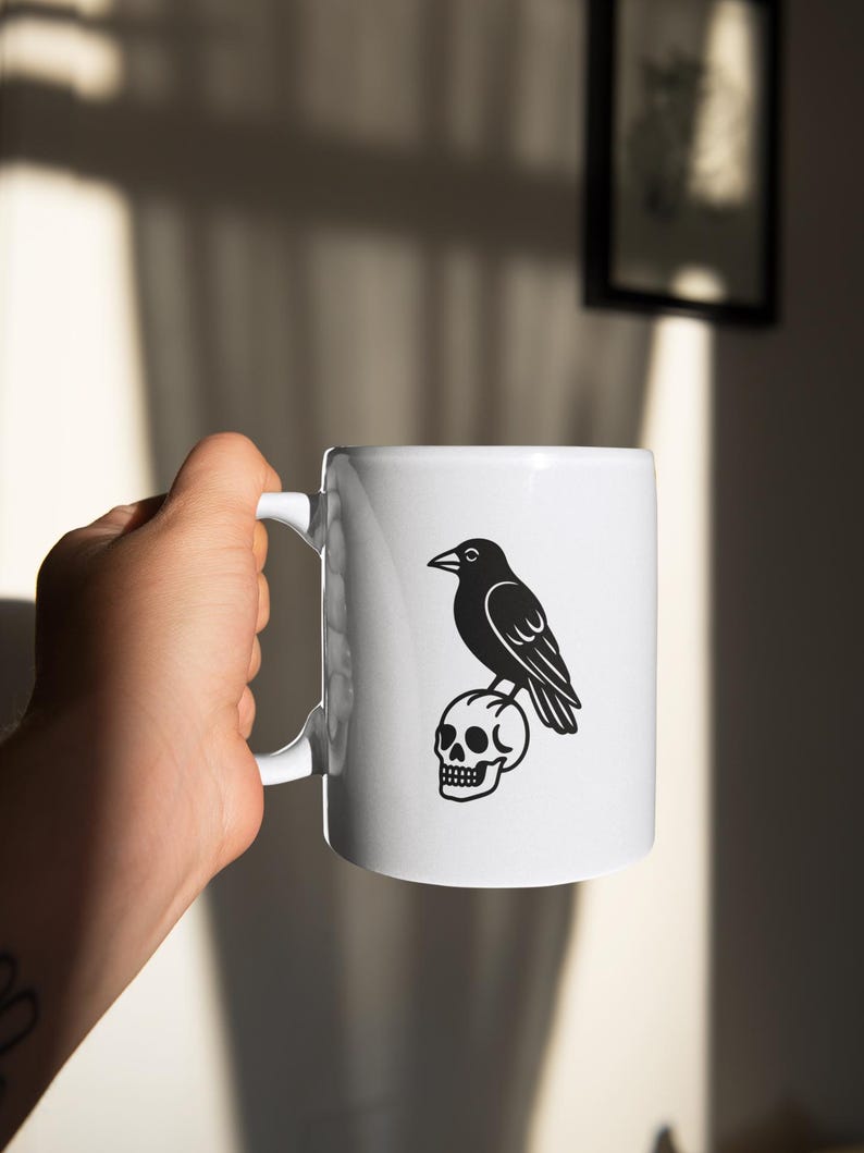 May include: White ceramic mug featuring a black illustration of a raven perched atop a skull. The mug has a curved handle and is held by a person. The design is a simple, yet striking, black and white graphic.