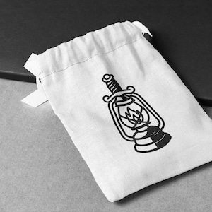 May include: A white drawstring bag with a black graphic of a vintage lantern with a sword through it. The lantern has a flame inside. The bag is on a gray surface.