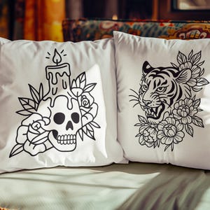 May include: Two white throw pillows with black graphic designs. One features a skull with a candle and floral accents, the other a tiger's face surrounded by flowers. The pillows are on a patterned sofa.