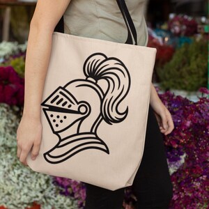 May include: Beige tote bag featuring a black line drawing of a knight's helmet with a visor and a flowing plume. The bag is carried by a person. The background includes colorful flowers.