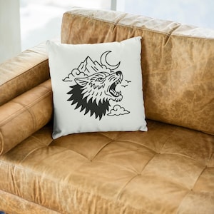 May include: White square throw pillow featuring a black illustration of a howling wolf, mountains, a crescent moon, and clouds. The pillow is placed on a brown leather couch.