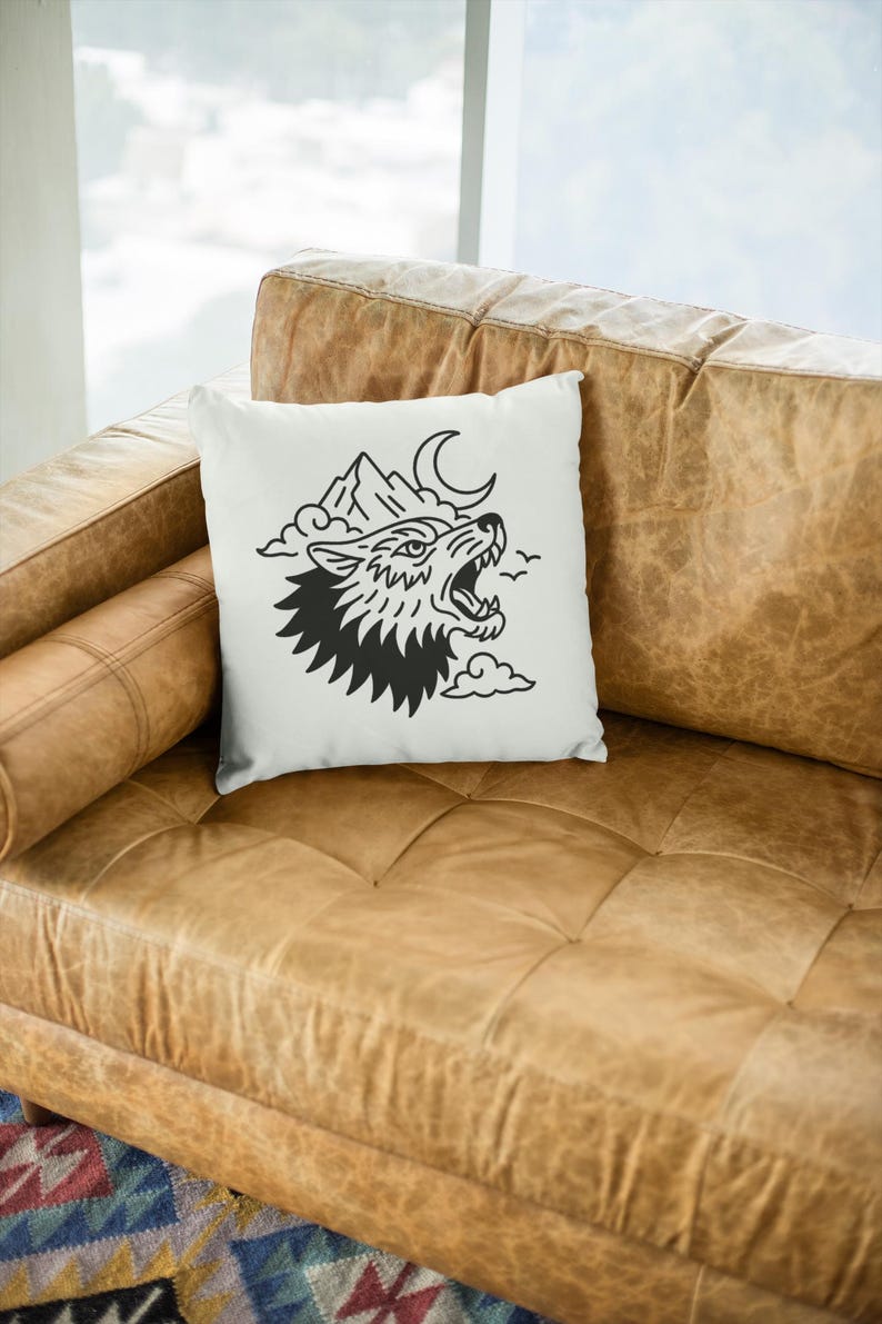 May include: White square throw pillow featuring a black illustration of a howling wolf, mountains, a crescent moon, and clouds. The pillow is placed on a brown leather couch.