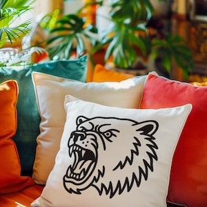 May include: A decorative pillow with a cream-colored background features a black illustration of a bear's head with an open mouth. The pillow is part of a collection of colorful pillows in shades of orange, teal, and red.