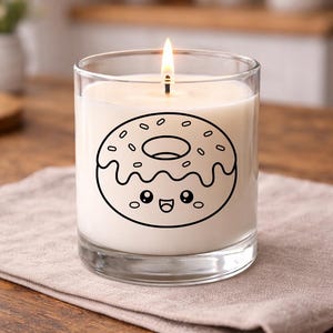 Puede incluir: Una vela encendida en un tarro de cristal transparente. La vela tiene un interior de cera blanca y un contorno negro de un donut con una cara sonriente. La vela est&aacute; sobre una servilleta de lino beige sobre una mesa de madera.