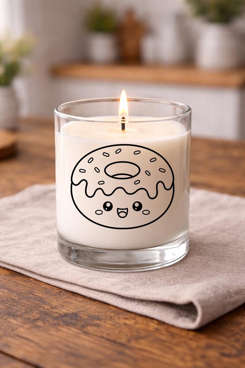 Puede incluir: Una vela encendida en un tarro de cristal transparente. La vela tiene un interior de cera blanca y un contorno negro de un donut con una cara sonriente. La vela est&aacute; sobre una servilleta de lino beige sobre una mesa de madera.
