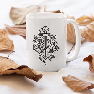 May include: White ceramic mug featuring a black line drawing of an anchor intertwined with roses and leaves. The mug has a curved handle and is surrounded by dried leaves on a white surface.
