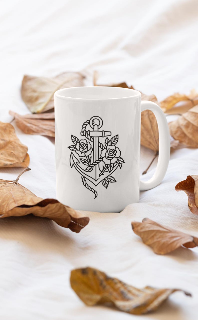 May include: White ceramic mug featuring a black line drawing of an anchor intertwined with roses and leaves. The mug has a curved handle and is surrounded by dried leaves on a white surface.