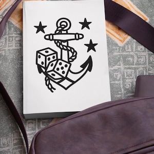 May include: A white notebook featuring a black anchor design with dice and stars. The anchor is intertwined with rope, and the dice show various numbers. The notebook is next to a brown shoulder bag with a dark strap.