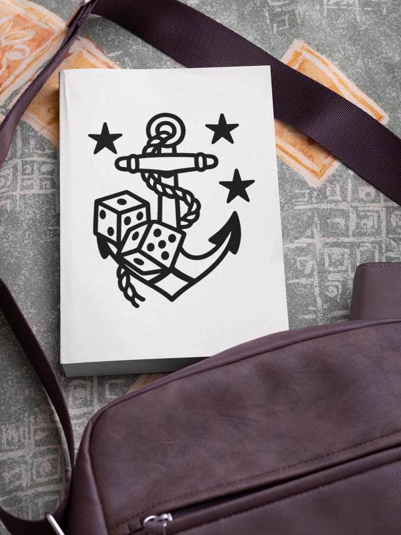 May include: A white notebook featuring a black anchor design with dice and stars. The anchor is intertwined with rope, and the dice show various numbers. The notebook is next to a brown shoulder bag with a dark strap.