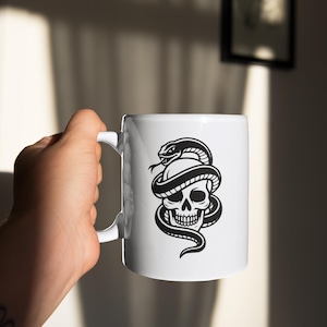 May include: White ceramic mug featuring a black and white illustration of a skull entwined by a snake. The mug has a standard handle and a glossy finish. The design is a bold, graphic style, suitable for beverages.