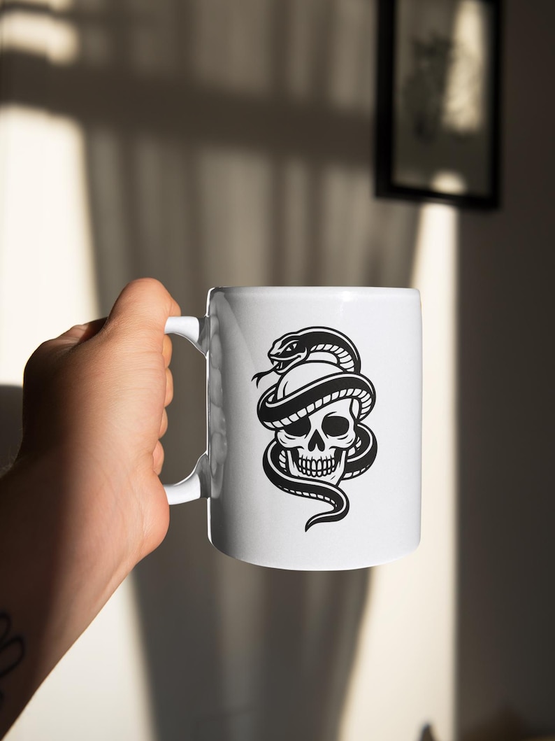 May include: White ceramic mug featuring a black and white illustration of a skull entwined by a snake. The mug has a standard handle and a glossy finish. The design is a bold, graphic style, suitable for beverages.