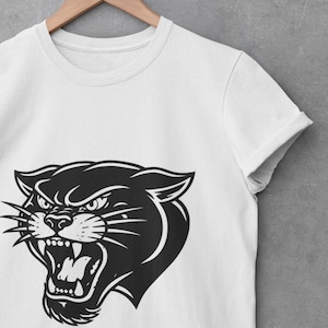 May include: White t-shirt featuring a black panther head graphic. The panther is depicted with an open mouth, bared teeth, and an angry expression. The shirt is hanging on a wooden hanger against a gray background.