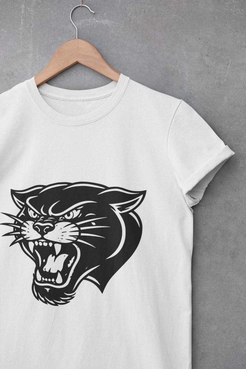 May include: White t-shirt featuring a black panther head graphic. The panther is depicted with an open mouth, bared teeth, and an angry expression. The shirt is hanging on a wooden hanger against a gray background.