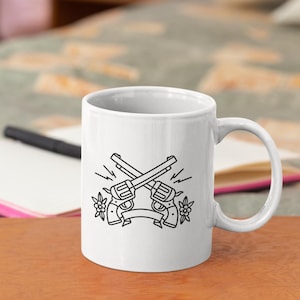 May include: White ceramic mug featuring a black and white illustration of two crossed revolvers with a banner and floral accents. The mug has a curved handle and is set on a wooden surface.