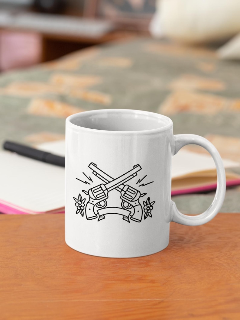 May include: White ceramic mug featuring a black and white illustration of two crossed revolvers with a banner and floral accents. The mug has a curved handle and is set on a wooden surface.