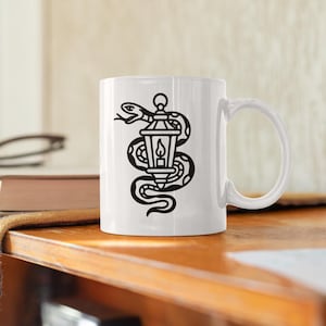 May include: White ceramic mug featuring a black illustration of a snake coiled around a lantern with a lit candle inside. The mug has a curved handle and is set on a wooden surface.
