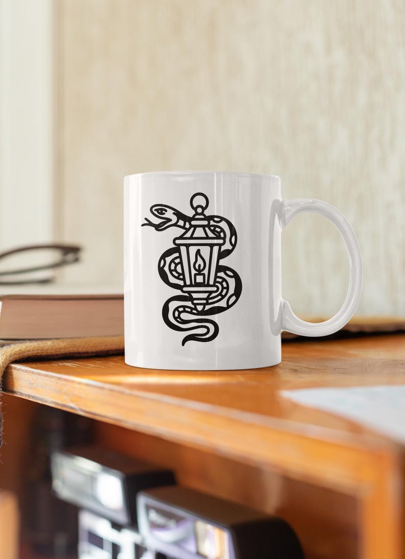 May include: White ceramic mug featuring a black illustration of a snake coiled around a lantern with a lit candle inside. The mug has a curved handle and is set on a wooden surface.