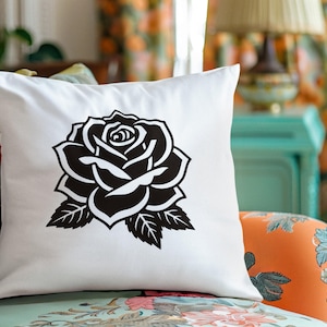 May include: White square throw pillow featuring a large black rose design. The rose is a detailed illustration with petals and leaves. The pillow is placed on a floral patterned chair, suggesting home decor.
