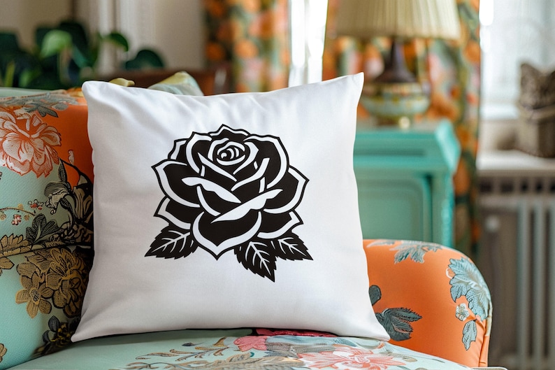 May include: White square throw pillow featuring a large black rose design. The rose is a detailed illustration with petals and leaves. The pillow is placed on a floral patterned chair, suggesting home decor.