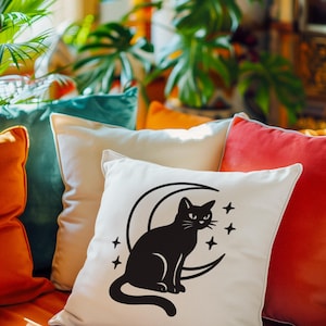 May include: A decorative pillow with a black cat silhouette sitting on a crescent moon, surrounded by four stars. The pillow is white and rests among other colorful pillows in shades of orange, teal, and red.