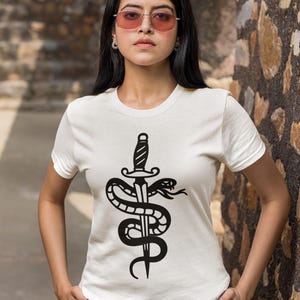 May include: White t-shirt featuring a black graphic of a dagger piercing a coiled snake. The design is centered on the front of the shirt. The model is wearing pink-tinted sunglasses and light-colored pants.