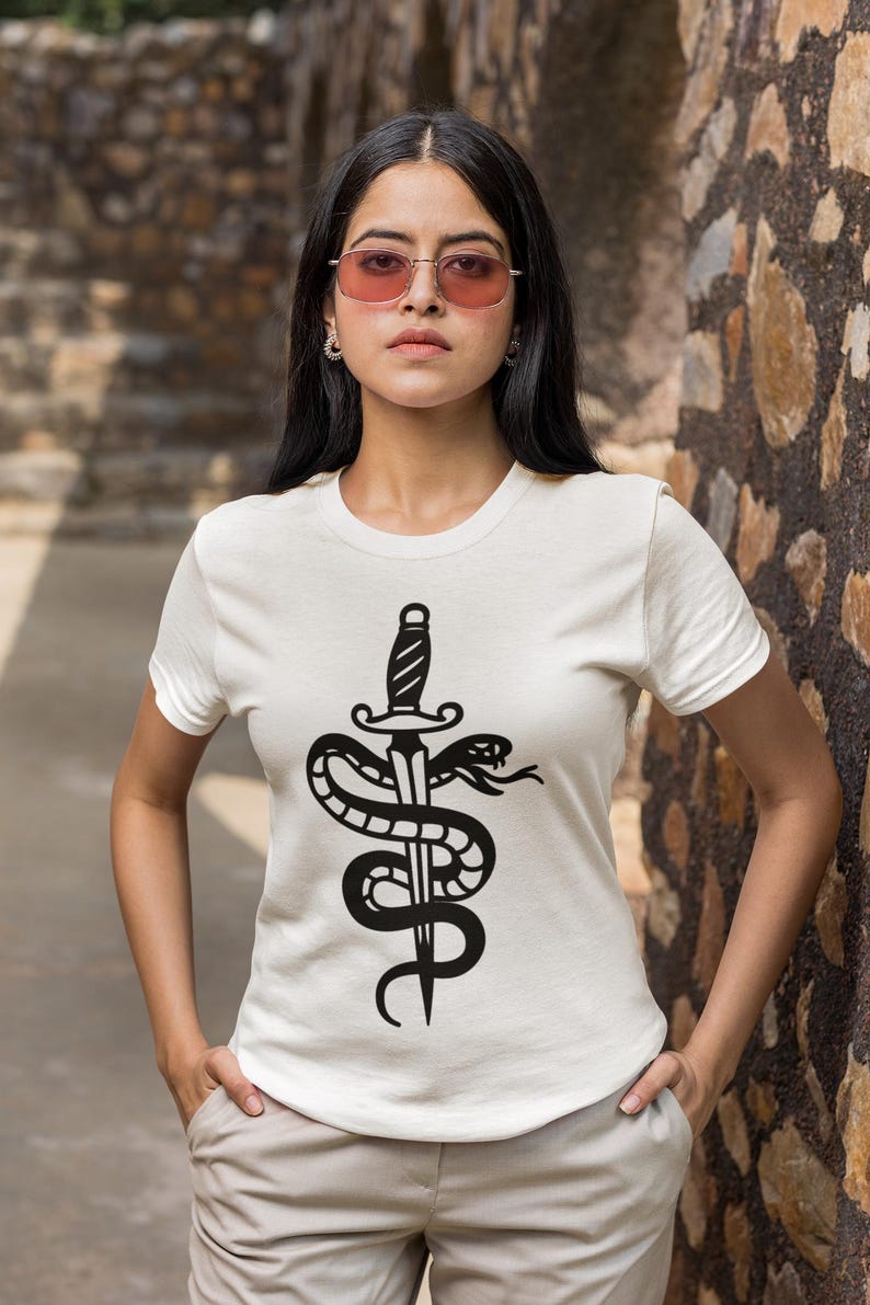 May include: White t-shirt featuring a black graphic of a dagger piercing a coiled snake. The design is centered on the front of the shirt. The model is wearing pink-tinted sunglasses and light-colored pants.