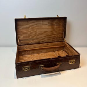 May include: Open brown leather briefcase with a crocodile pattern. The interior is lined with wood and a lighter brown fabric. The briefcase has a brown leather handle and gold-coloured latches.