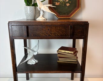 Puede incluir: Una mesa auxiliar de madera marrón oscuro con un cajón, que incluye una orquídea blanca en una maceta moteada, dos figuras de pájaros de cerámica blanca, un cuadro floral octogonal con marco rojo y una pila de cinco libros con bordes dorados.