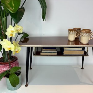 May include: A rectangular coffee table with a dark brown top, a gold-toned metal trim, and four tapered black legs. The table features an integrated lower shelf holding several books, and the top surface holds three ceramic mugs with brown patterns.