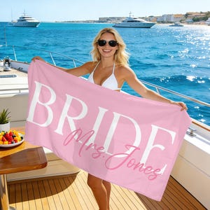 Puede incluir: Una toalla de playa rosa con la palabra "BRIDE" en letras blancas grandes y "Mrs. Jones" en cursiva. La toalla se sostiene en un barco con el océano y otros barcos al fondo.