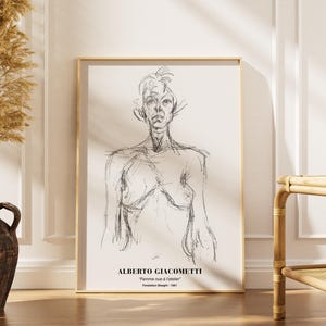 May include: A framed sketch of a figure by Alberto Giacometti, titled "Femme nue à l'atelier." The artwork is in a gold frame and is displayed against a white wall. A decorative vase with dried plants is to the left.