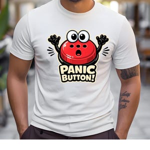 Op de afbeelding: Wit T-shirt met een cartoon rode paniekknop karakter met zwarte armen en grote ogen. De tekst "PANIC BUTTON!" staat onder het karakter in een gele en witte lettertype. Het T-shirt heeft een ronde hals.