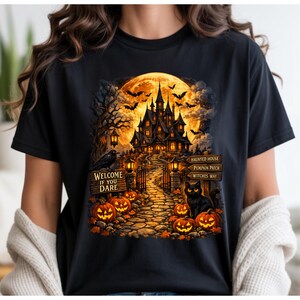 Puede incluir: Una camiseta negra con un gráfico detallado de una casa encantada bajo una luna llena, un camino de piedra, calabazas, un gato negro y murciélagos. El texto dice: WELCOME IF YOU DARE, HAUNTED HOUSE, PUMPKIN PATCH y WITCHES WAY.