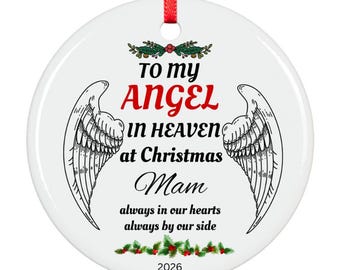 Op de afbeelding: Een ronde witte keramische kerstornament met een rood lint. Het bevat zwarte en rode tekst met de tekst To my ANGEL IN HEAVEN at Christmas Mam always in our hearts always by our side 2026, geflankeerd door twee engelenvleugels en hulst.