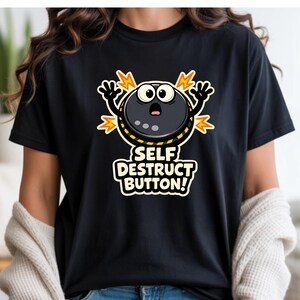 May include: Black t-shirt featuring a cartoon self-destruct button design. The graphic includes a round, black button with eyes, arms, and a surprised expression, surrounded by yellow and orange explosion lines. The text "SELF DESTRUCT BUTTON!" is below.