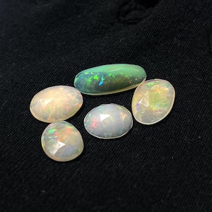 May include: Five loose opal gemstones with faceted surfaces. One stone is elongated and dark green with iridescent flashes, while the other four are oval-shaped, translucent white stones displaying a play of color with red, blue, and green hues.
