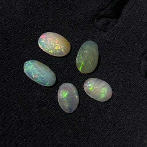 May include: Five oval-shaped opals with a translucent, milky white base color. Each gemstone displays a distinct play-of-color, featuring flashes of green, yellow, and red light across their smooth, polished surfaces against a dark background.