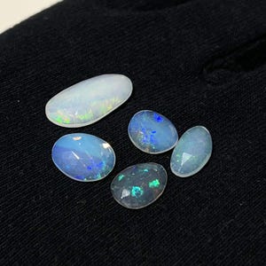 May include: Four oval-shaped cabochon opals with faceted surfaces. The largest opal is translucent white with green and yellow flashes. The three smaller opals display blue, green, and teal iridescent play-of-color against a semi-translucent base.