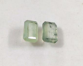 May include: Two rectangular, emerald-cut gemstones with a pale mint green hue. Each stone features a faceted surface that reflects light, and the right stone displays visible dark, needle-like inclusions throughout its translucent body.