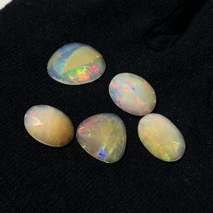 May include: Five loose, faceted opal gemstones with a translucent, milky white base and iridescent flashes of red, green, and blue color play. The stones are arranged in a cluster, each displaying a smooth, polished surface and a unique oval shape.